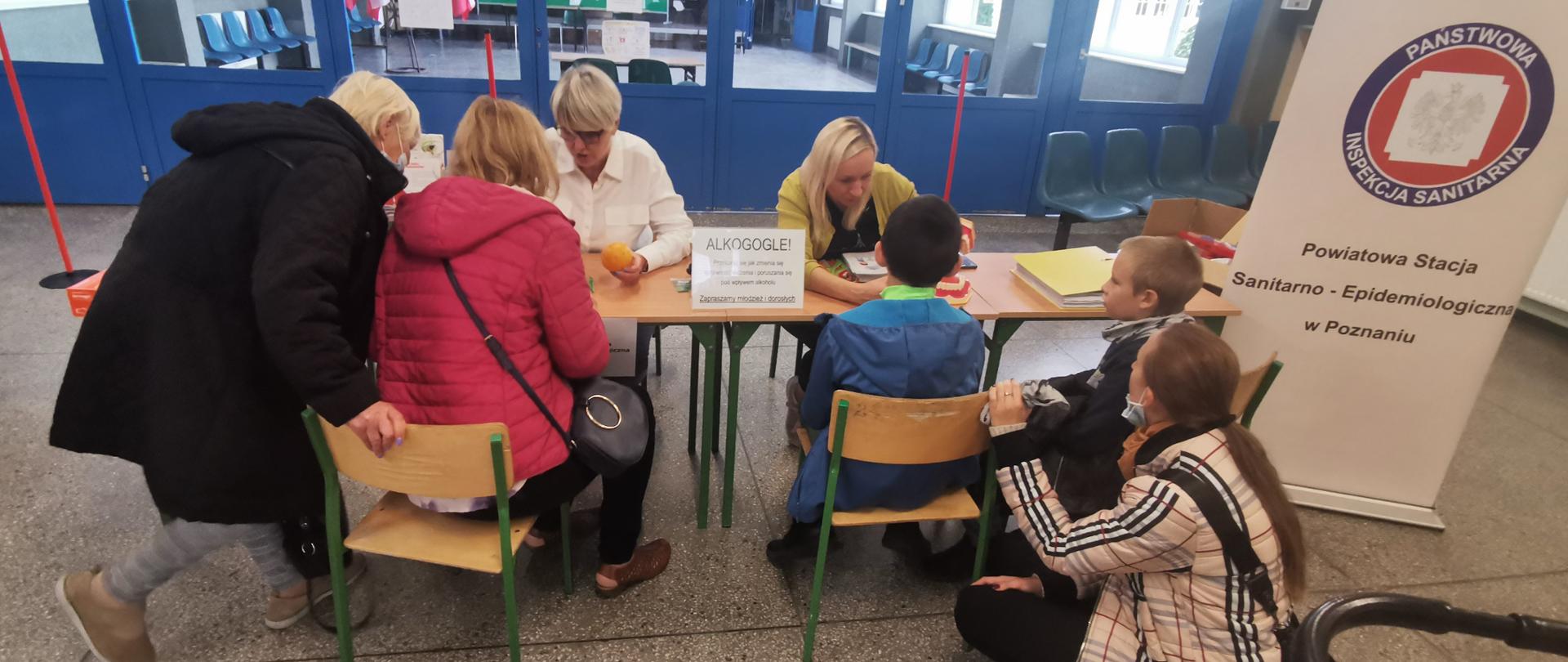 Przy stoisku PSSE, po lewej stronie dwie osoby dorosłe uczestniczą w instruktarzu prawidłowego usuwania kleszcza, po prawej dzieci uczestniczą w instruktarzu prawidłowego mycia zębów.