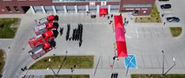 Na zdjęciu widoczny plac wewnętrzny komendy w trakcie uroczystego podniesienie flagi państwowej. Na pierwszym planie poczet flagowy. Za pocztem z lewej strony zgromadzeni goście pod czerwonym namiotem, z prawej strony strażacy ustawieni w rzędach. Za strażakami ustawione samochody gaśnicze. W tle budynek komendy. 