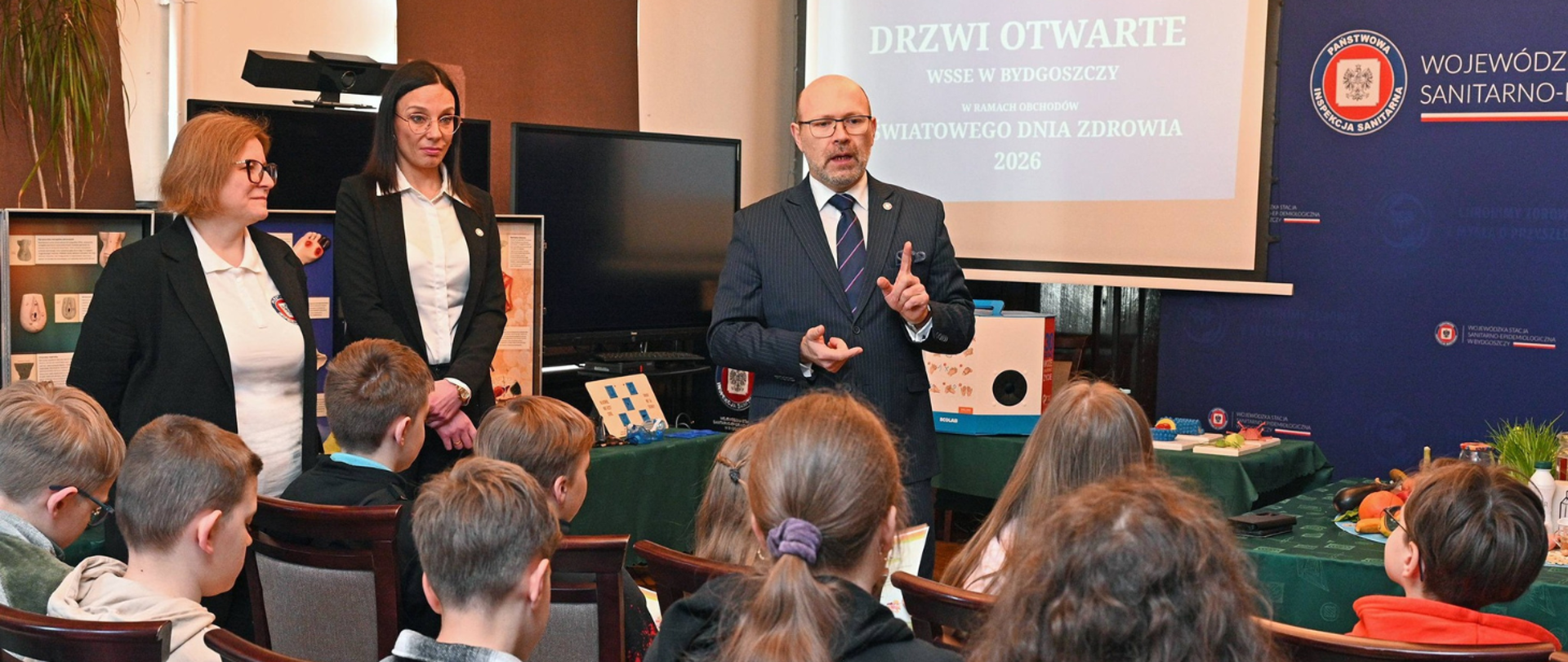 Państwowy Wojewódzki Inspektor Sanitarny przemawia do grupy dzieci podczas Drzwi Otwartych w WSSE w Bydgoszczy; obok stoją dwie pracownice, a w tle widoczna jest prezentacja oraz stanowiska edukacyjne.