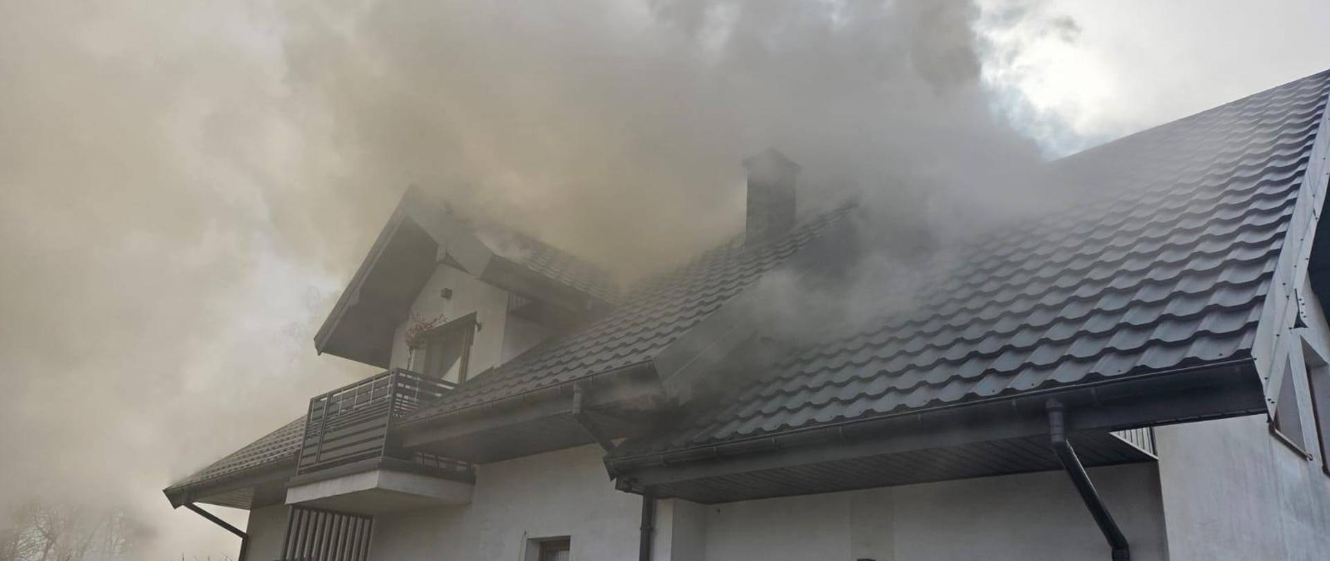 Gęsty dym wydobywa się z dachu domu jednorodzinnego pokrytego blachodachówką.