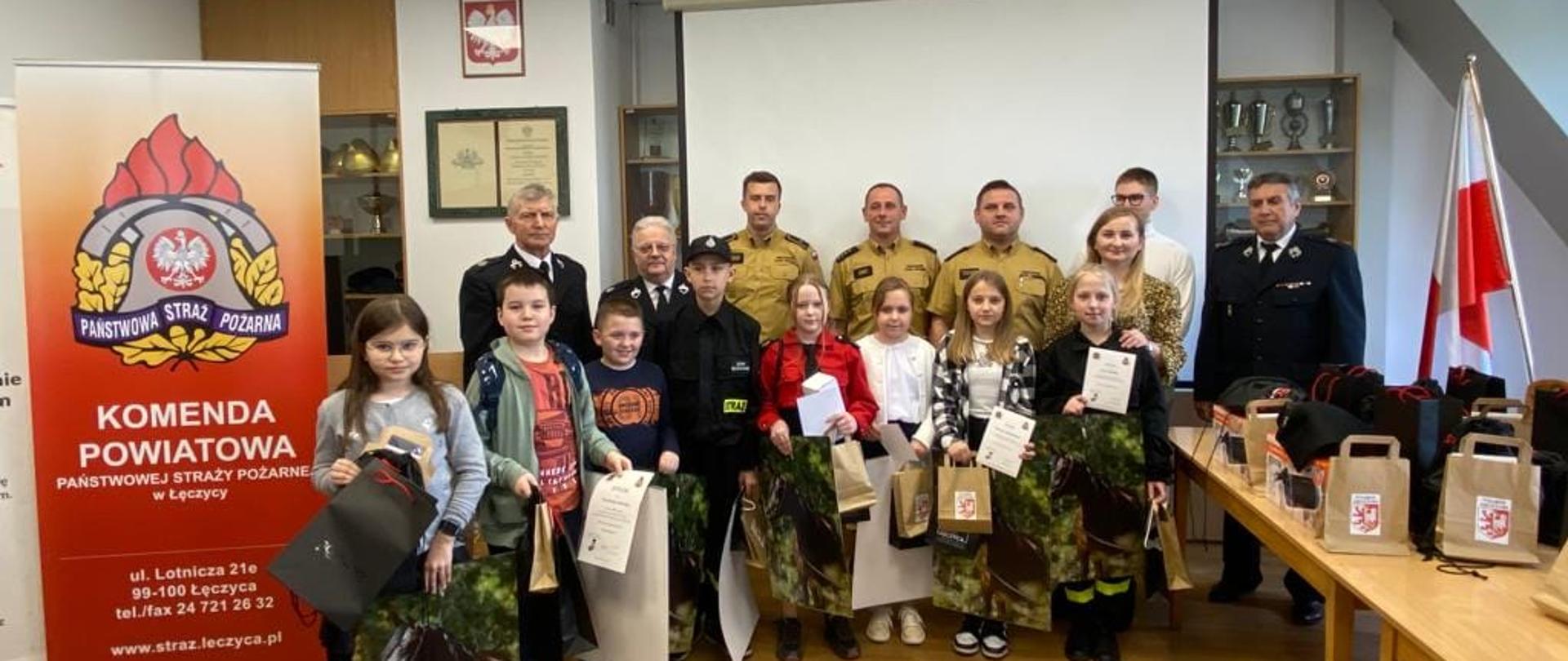 Na zdjęciu widać trzech strażaków w żółtych strojach, trzech w ciemnych strojach, kobietę oraz mężczyznę oraz dzieci, które trzymają w rękach nagrody. Wszyscy pozują do zdjęcia. W tle widać godło i flagę Polski, obrazy, baner z logiem Państwowej Straży Pożarnej oraz danymi teleadresowymi Komendy Powiatowej PSP w Łęczycy oraz szafę z szybą za którą znajdują się zabytkowe hełmy oraz puchary. Na stole stoją czarne i szare torby z nagrodami. Wydarzenie odbywa się wewnątrz budynku. Jest widno.