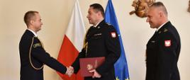 Na zdjęciu znajduje się trzech strażaków w mundurach wyjściowych, jeden z lewej i dwóch z prawej stoją na przeciwko siebie, dwóch strażaków stoi blisko siebie i podają sobie teczkę z dokumentem, tle flagi polski i unii europejskiej