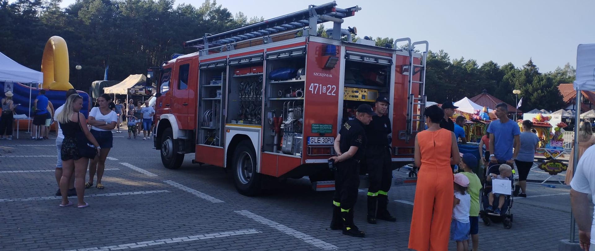 Na zdjęciu widoczny pojazd ratowniczo-gaśniczy, przy którym stoją strażacy pokazjujący wyposażenie pojazdu uczestnikom pikniku.