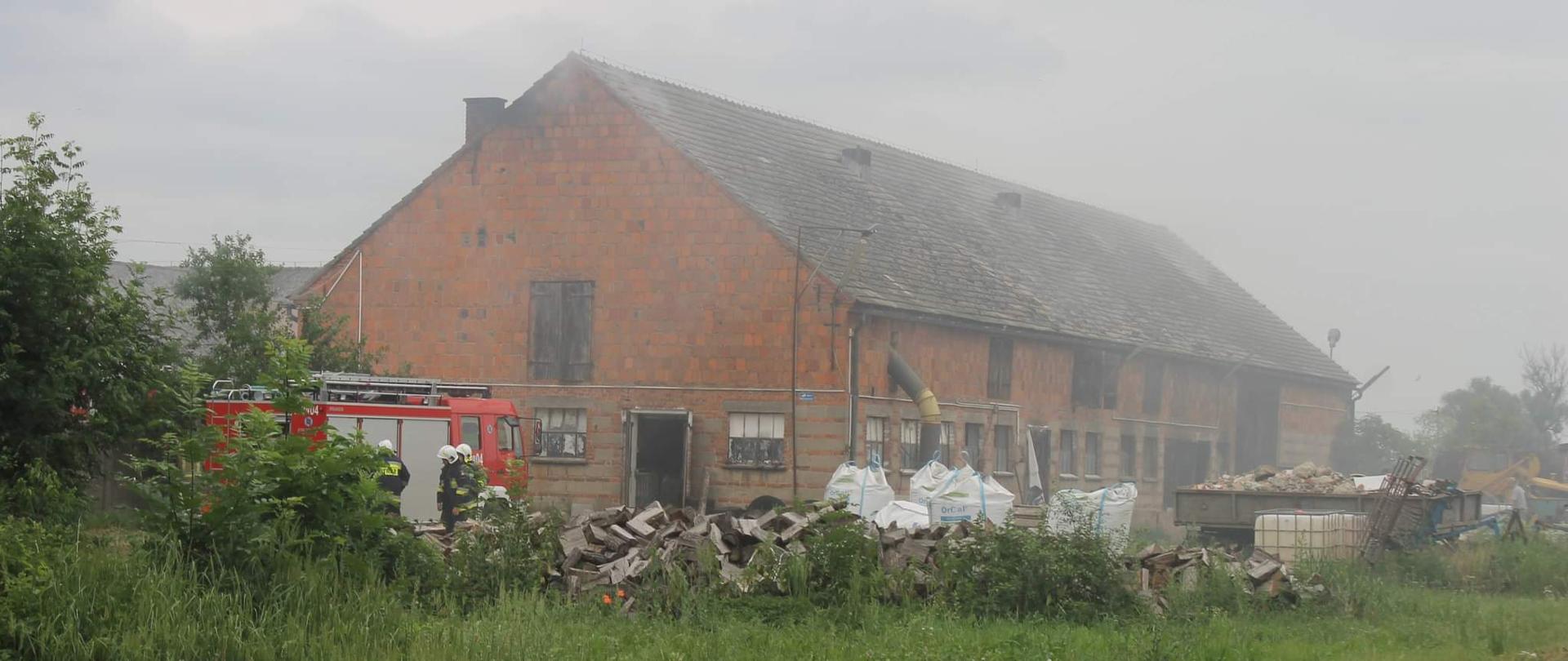 Pożar kotłowni w budynku gospodarczym. Przed obiektem stoi samochód pożarniczy. Obok strażacy OSP prowadzący działania gaśnicze. Przed pojazdem widoczne wory z materiałem opałowym. Trwają działania gaśnicze. Obok budynku duża ilość maszyn rolniczych oraz materiałów różnego pochodzenia.