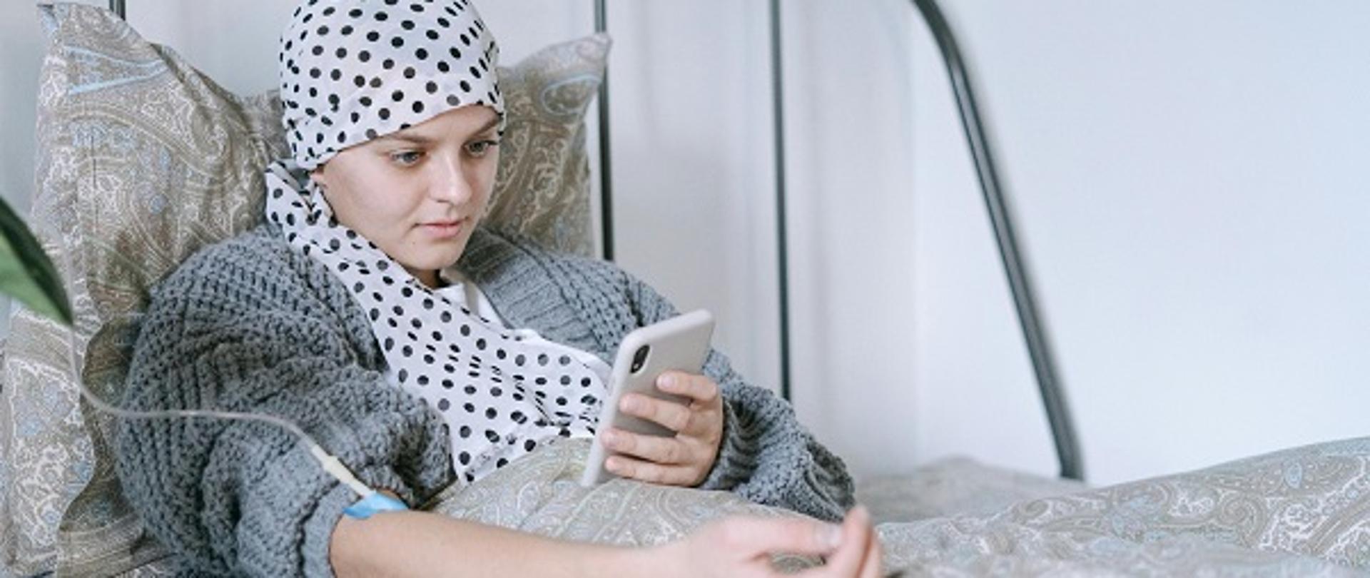 pacjentka w chustce na głowie, leżąca w łóżku, przyjmuje kroplówkę dożylnie, drugą ręką przegląda treści w telefonie