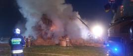 Zdjęcie zadymionych balotów słomy, w centralnym miejscu widoczne płomienie. Trzech strażaków w umundurowaniu specjalnym, w hełmach podaje prądy wody na stóg słomy. Po lewej stronie strażak w umundurowaniu specjalnym, w hełmie stojący tyłem. Po prawej stronie przednia część pojazdu pożarniczego z włączonymi światłami błyskowymi koloru niebieskiego.