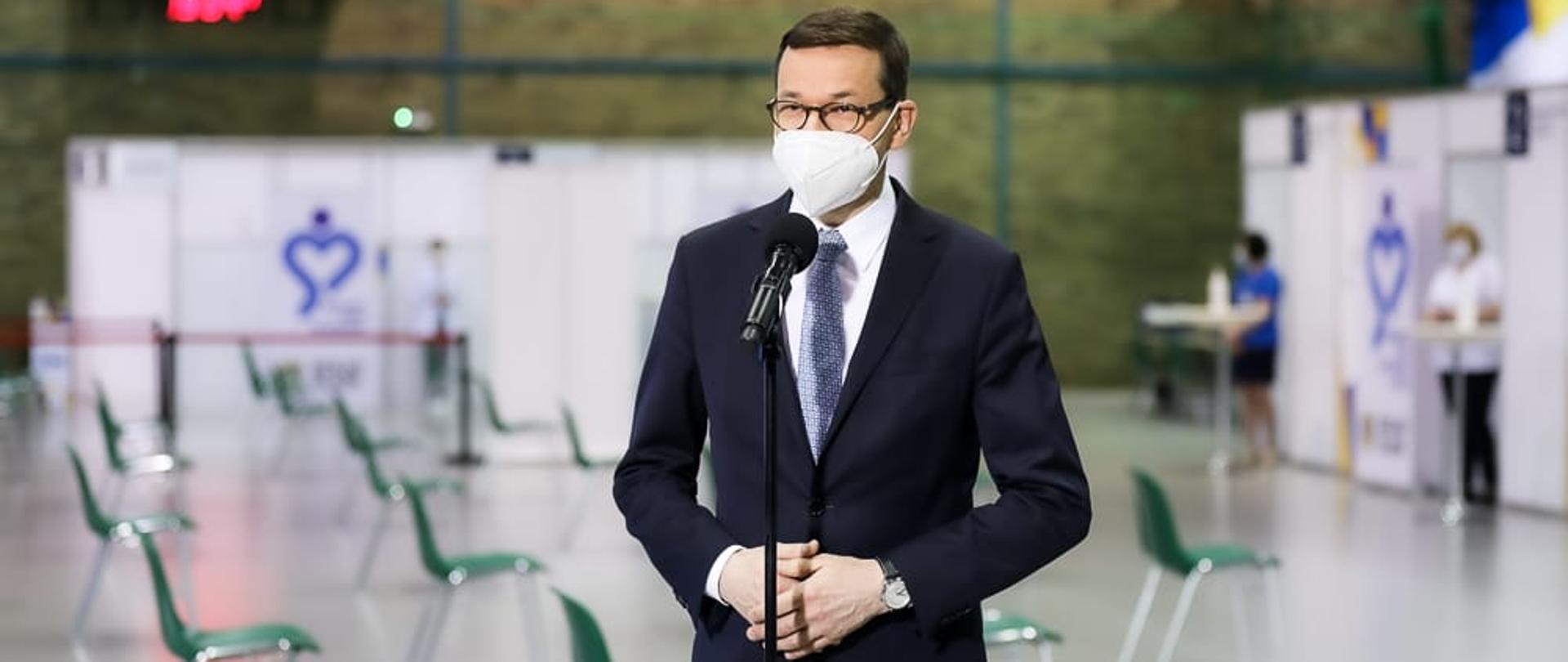 Premier Mateusz Morawiecki stoi przy mikrofonie. W tle Punkt Szczepień Powszechnych zorganizowany w hali sportowej.