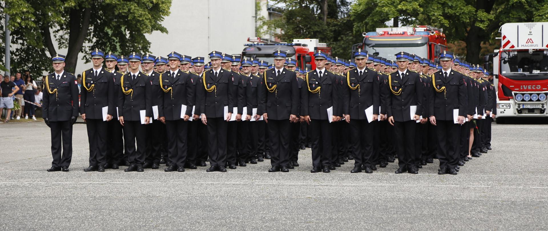 Grupa strażaków w mundurach galowych stoi na placu podczas promocji