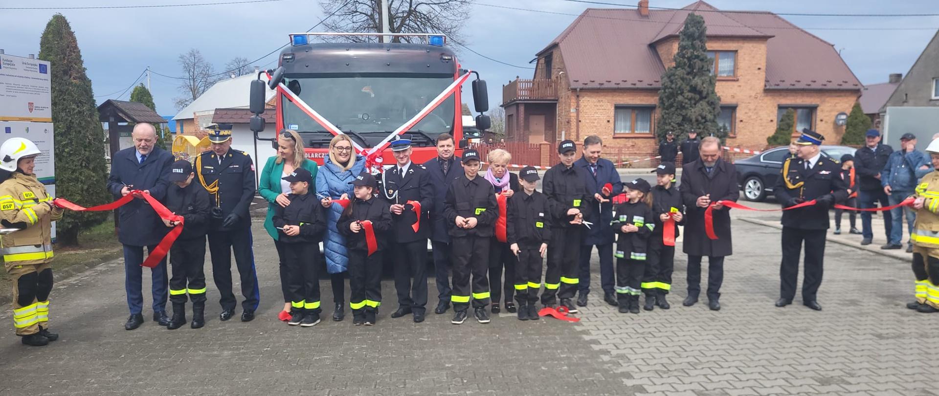 Zaproszeni goście wraz z młodzieżową drużyną pożarniczą stojący przed samochodem strażackim i przecinający czerwoną wstęgę. W tle zabudowania i inni strażacy.
