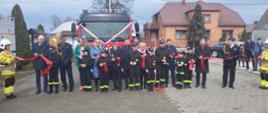 Zaproszeni goście wraz z młodzieżową drużyną pożarniczą stojący przed samochodem strażackim i przecinający czerwoną wstęgę. W tle zabudowania i inni strażacy.