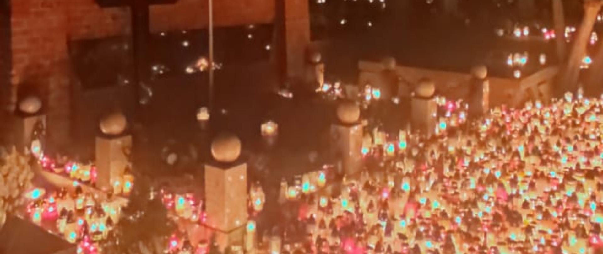 Zdjęcie przedstawia różnokolorowe zapalone znicze ustawione obok siebie na cmentarzu. Znicze różnych wielkości, w kolorach białym, czerwonym oraz żółtym. Zdjęcie wykonane w porze nocnej.