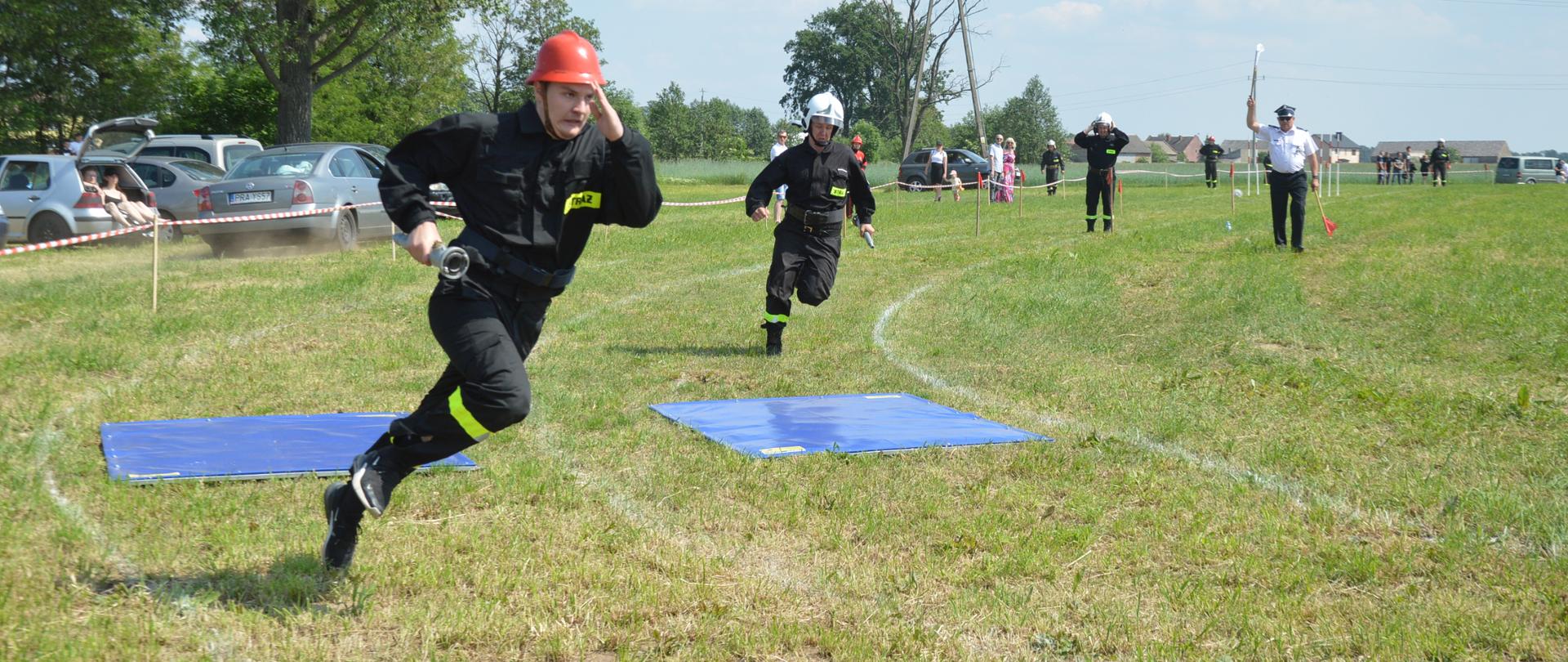 Zawody sportowo-pożarnicze w Zawadach. Na placu ćwiczeń trwa konkurencja "sztafeta pożarnicza 7x50". Startują dwie drużyny męskie. zawodnicy przeskakują przez niebieski "rów" . W tle inni zawodnicy, sędzia, kibice oraz samochody..