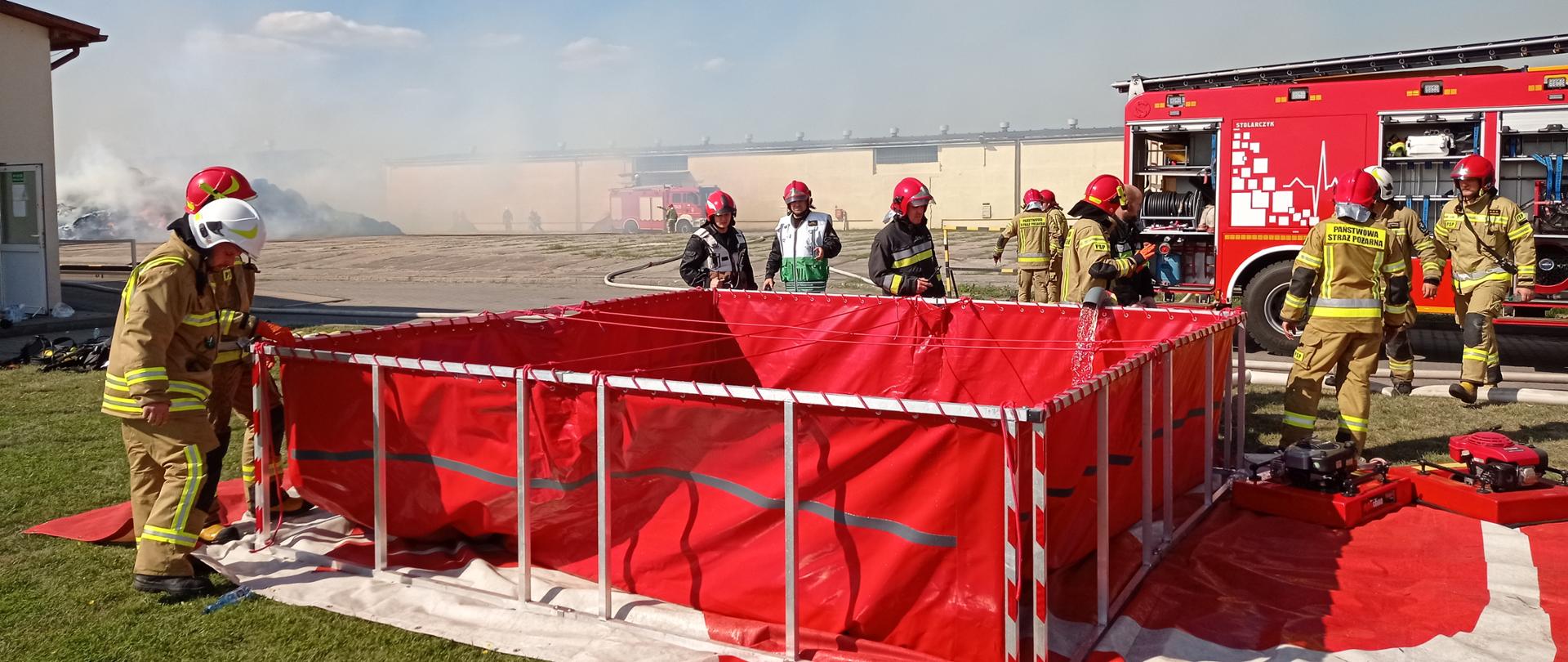 Zdjęcie przedstawia strażaków gaszących pożar w zakładzie produkującym biomasę, którzy sprawiają przenośny zbiornik brezentowy na wodę i uzupełniają wodę wężami pożarniczymi.
W tle budynki i palące się stogi słomy.
