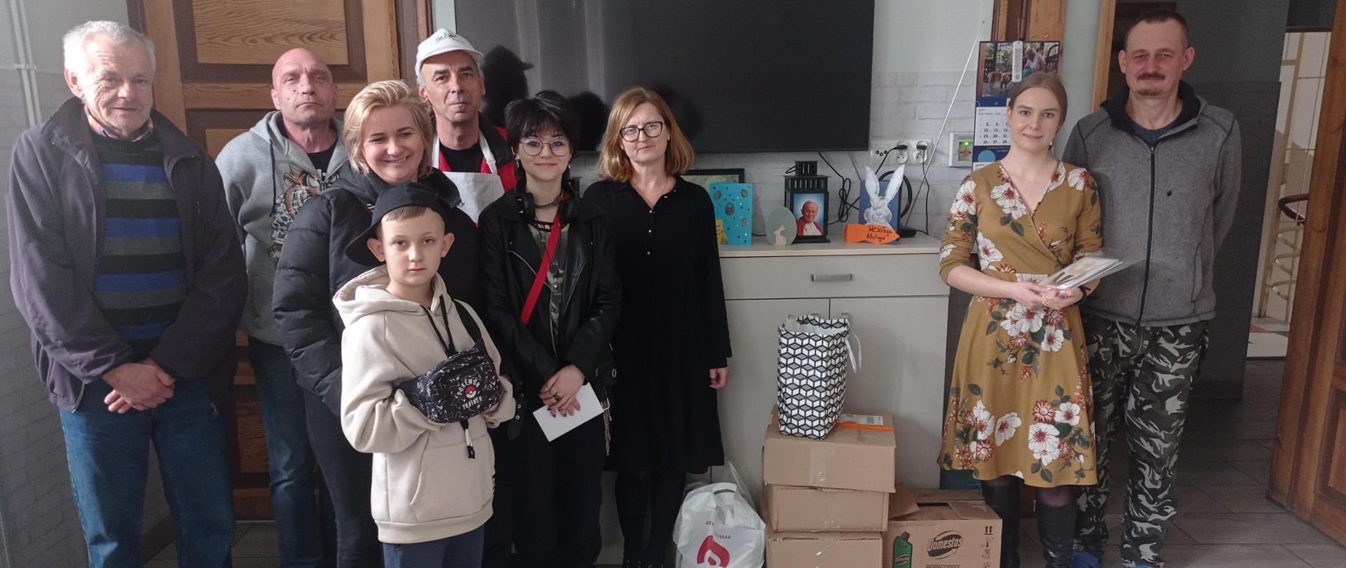 Grupa osób – kilkoro starszych, nastolatka i chłopiec – w pomieszczeniu gospodarczym. Stoją, patrzą w stronę robiącego zdjęcie. Na ziemi stoją kartony i siatki