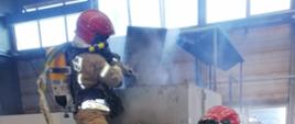 Strażacy wyposażeni w środki ochrony indywidualnej, w tym aparaty ochrony układu oddechowego podczas prac związanych z gaszeniem pianą maszyny w hali zakładu.
