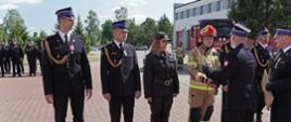 Komendant powiatowy w łowiczu oraz jego zastępca wręczają pamiątkowe dyplomy z okazji jubileuszu trzydziestolecia PSP strażakom, w tle budynek garażowy jednostki ratowniczo-gaśniczej oraz strażacy stojący w dwuszeregu, pogoda słoneczna