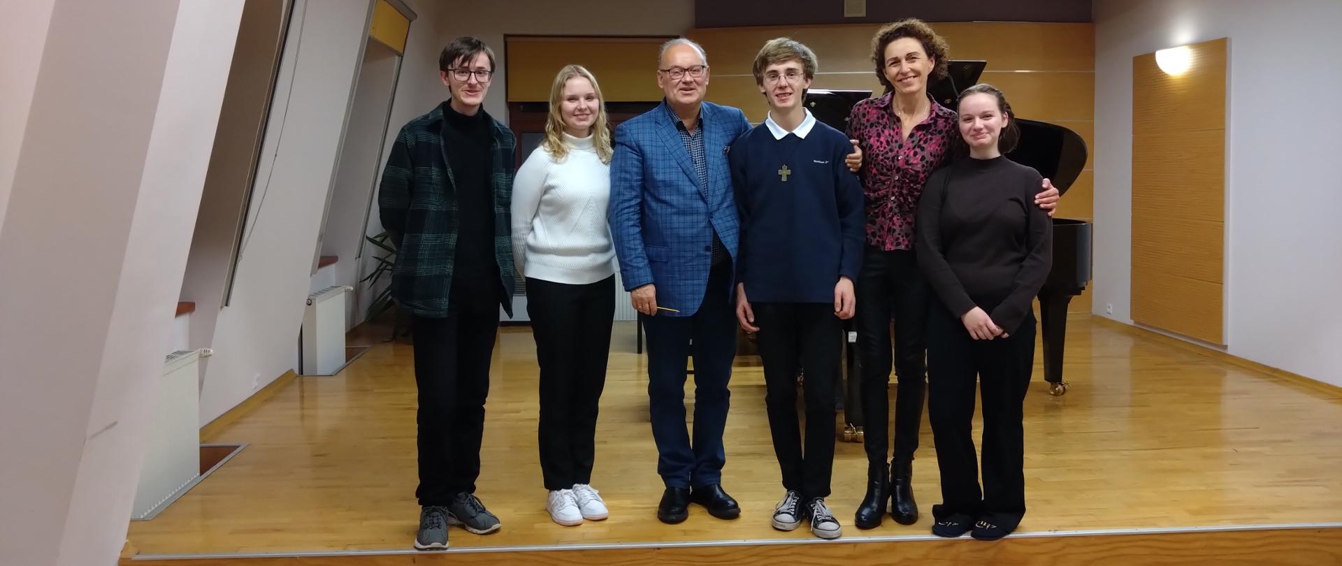 Uczniowie wraz z nauczycielem i prowadzącym warsztaty- Panem Arturem Jaroniem stoją w jednym rzędzie na scenie sali koncertowej. Za nimi dwa fortepiany, trzy okna przysłonięte roletą. Pod oknami grzejniki oraz nawilżacz powietrza. Nad nimi na suficie zapalone lampy sufitowe.