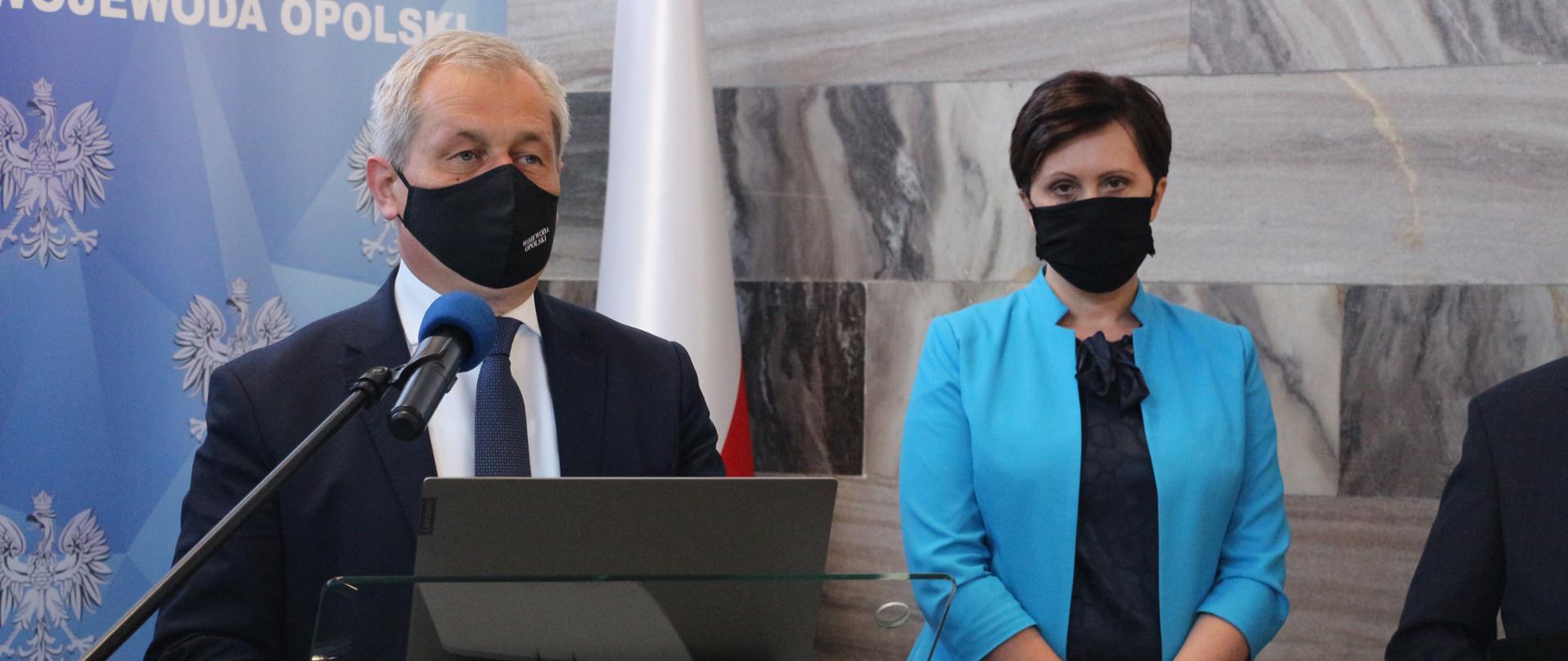 Na zdjęciu Wojewoda opolski i dyrektor opolskiego ZUS. Wojewoda stoi za szklaną mównicą, przed nim mikrofon na statywie. Na mównicy laptop. Za plecami wojewody stoi roll up. Obok wojewody po prawej stornie kadru dyrektorka ZUS w Opolu. Kobieta ubrana w granatowy żakiet i czarne spodnie. Spogląda w dół. Po prawej stronie kadru widać część tułowia pozostałej osoby.