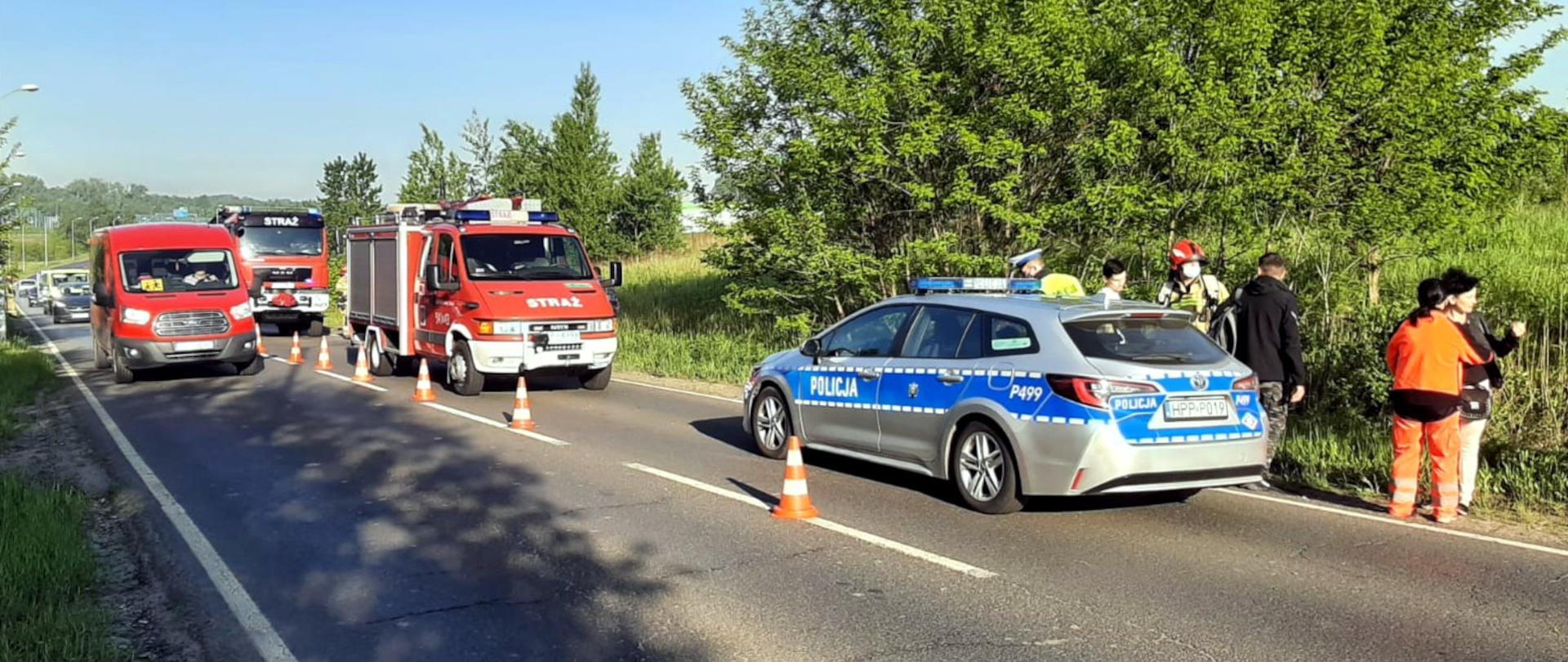 Zdjęcie przedstawia radiowóz policyjny, lekki samochód ratownictwa technicznego oraz średni samochód ratowniczo - gaśniczy stojące na poboczu drogi. Obok stoją ratownicy oraz uczestnicy wypadku.