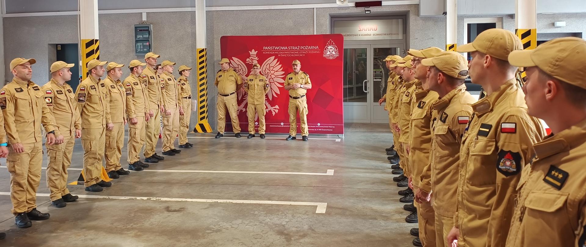 Zdjęcie przedstawia oficjalną zbiórkę strażaków wewnątrz przestronnego garażu jednostki ratowniczo-gaśniczej. Zgromadzeni funkcjonariusze są ubrani w jednolite, piaskowe mundury służbowe oraz czapki z daszkiem w tym samym kolorze. Stoją oni w dwóch równoległych szeregach naprzeciwko siebie, tworząc szpaler.
W głębi pomieszczenia, pomiędzy dwoma kolumnami oznaczonymi żółto-czarnymi pasami ostrzegawczymi, ustawiono czerwoną ściankę reklamową z godłem Polski oraz napisem „PAŃSTWOWA STRAŻ POŻARNA”. Przed ścianką stoi trzech mężczyzn w takich samych mundurach, z których jeden, znajdujący się pośrodku, przemawia do zebranych strażaków. Całe pomieszczenie jest oświetlone jasnym, górnym światłem, a na betonowej podłodze widoczne są białe linie wyznaczające miejsca postojowe.
