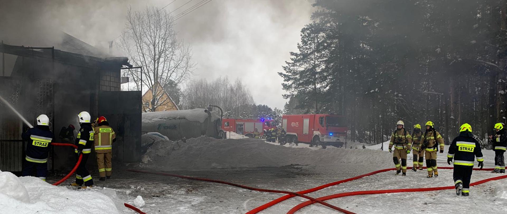 Na zdjęciu widać w tle samochody strażackie; strażacy podają prądy wody do wnętrza budynku