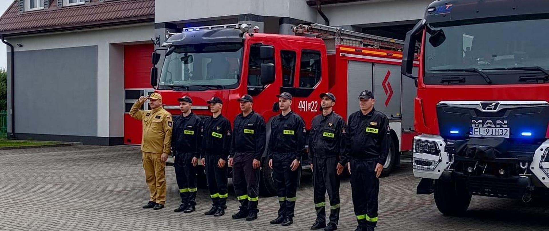 Na zdjęciu widać siedmiu strażaków stojących w jednym rzędzie przed samochodami strażackimi zaparkowanymi na placu przed budynkiem komendy. Jeden strażak ubrany jest w żółty strój a reszta w czarny. Wszyscy mają na głowach czapki. Jest widno.