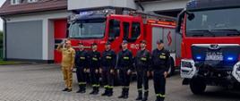 Na zdjęciu widać siedmiu strażaków stojących w jednym rzędzie przed samochodami strażackimi zaparkowanymi na placu przed budynkiem komendy. Jeden strażak ubrany jest w żółty strój a reszta w czarny. Wszyscy mają na głowach czapki. Jest widno.