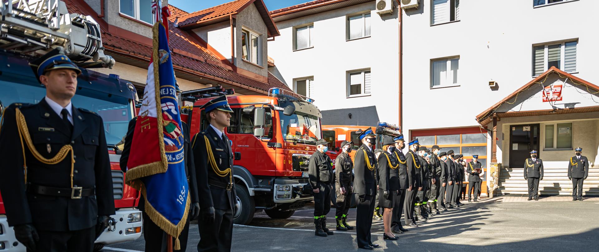 Zdjęcie przedstawia plac Komendy Powiatowej PSP w Ustrzykach Dolnych na którym odbywa się uroczysty apel z okazji dnia strażaka. Na placu w szyku ustawieni są strażacy w mundurach galowych, koszarowych i służbowych. Na pierwszym planie widać poczet sztandarowy oraz wozy bojowe. W tle widać komendanta powiatowego i jego zastępce którzy stoją przy wejściu głównym do Komendy.