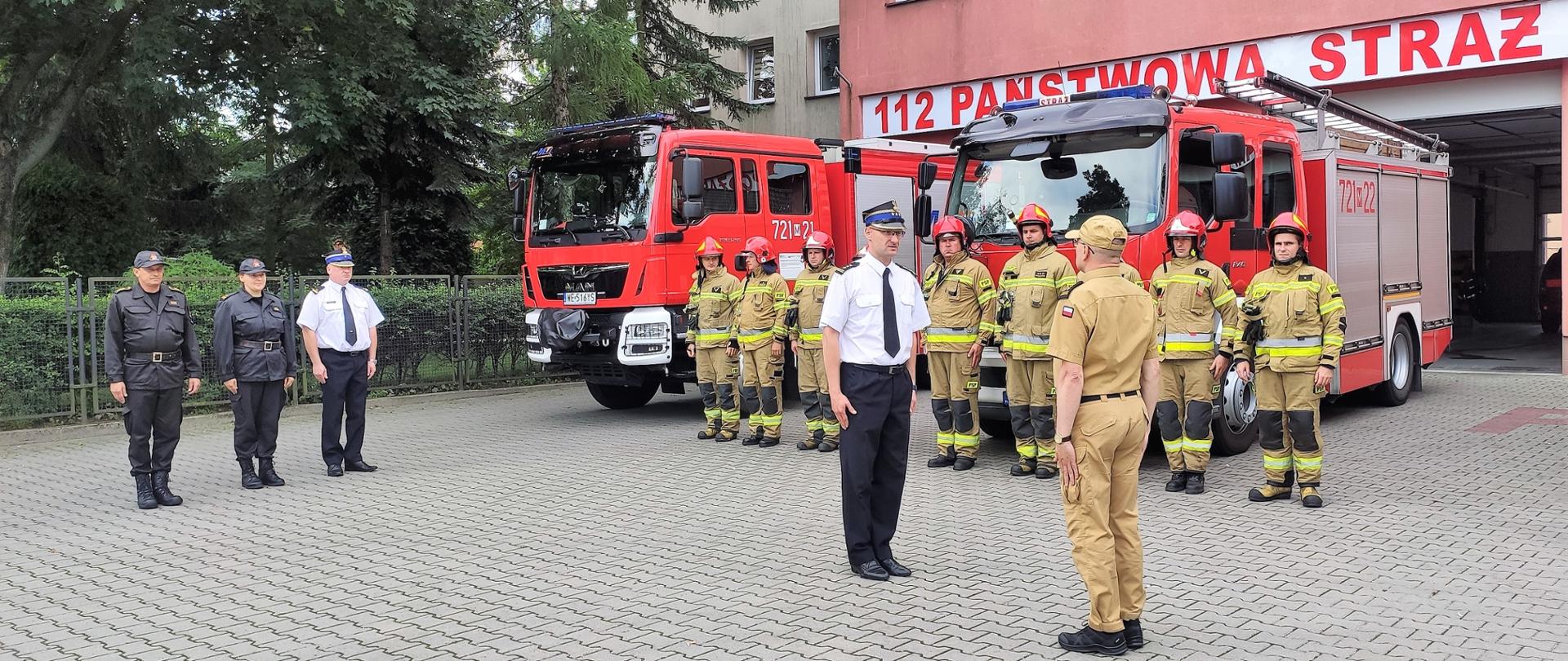 Komendant powiatowy PSP w Żyrardowie st. bryg. Mariusz Tymoszewicz składa meldunek zastępcy mazowieckiego komendanta wojewódzkiego PSP st. bryg. Dariuszowi Stumskiemu. W tle stoją strażacy i samochody strażackie.