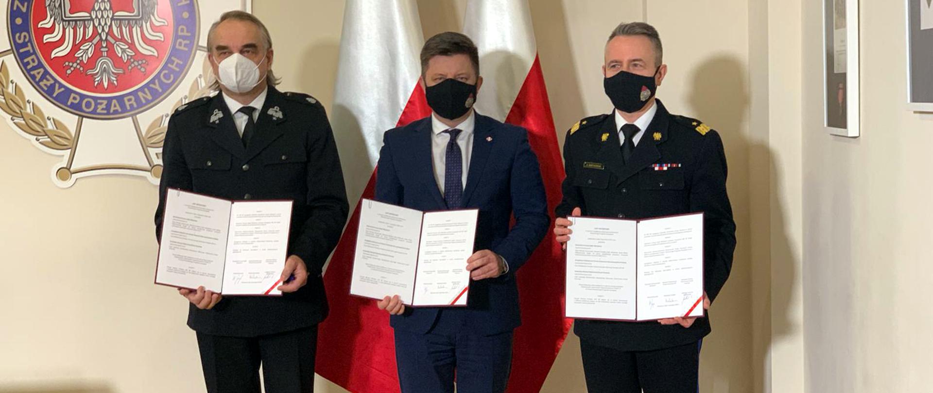 Postać w garniturze obok dwóch strażaków w mundurach galowych, wszyscy prezentują otwarte dokumenty