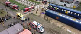 Widok z drona na miejsce przeprowadzanych ćwiczeń. Po lewej dwie karetki oraz cztery wozy bojowe PSP, po prawej przewrócony wagon pociągu oraz samochód osobowy, który zderzył się z pociągiem