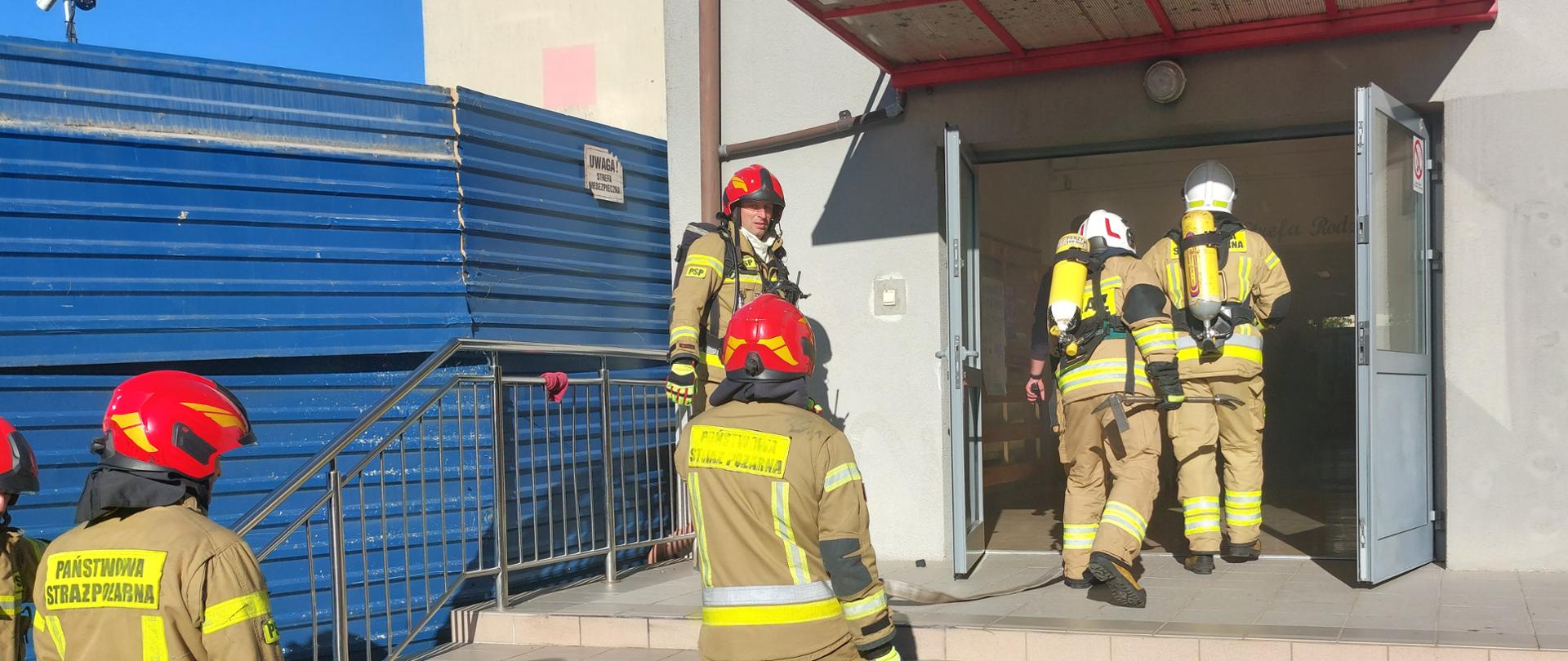 Na zdjęciu kilku strażaków w pełnym umundurowaniu ochronnym i czerwonych hełmach uczestniczy w ćwiczeniach przed wejściem do budynku szkoły. Dwóch z nich wchodzi do środka przez otwarte drzwi, a pozostali stoją na schodach i w pobliżu, przygotowując węże gaśnicze. Na ziemi rozłożony jest sprzęt strażacki, w tym rozdzielacz wodny i zwinięte węże. Scena odbywa się w słoneczny dzień przed budynkiem szkoły z czerwonym zadaszeniem nad wejściem.