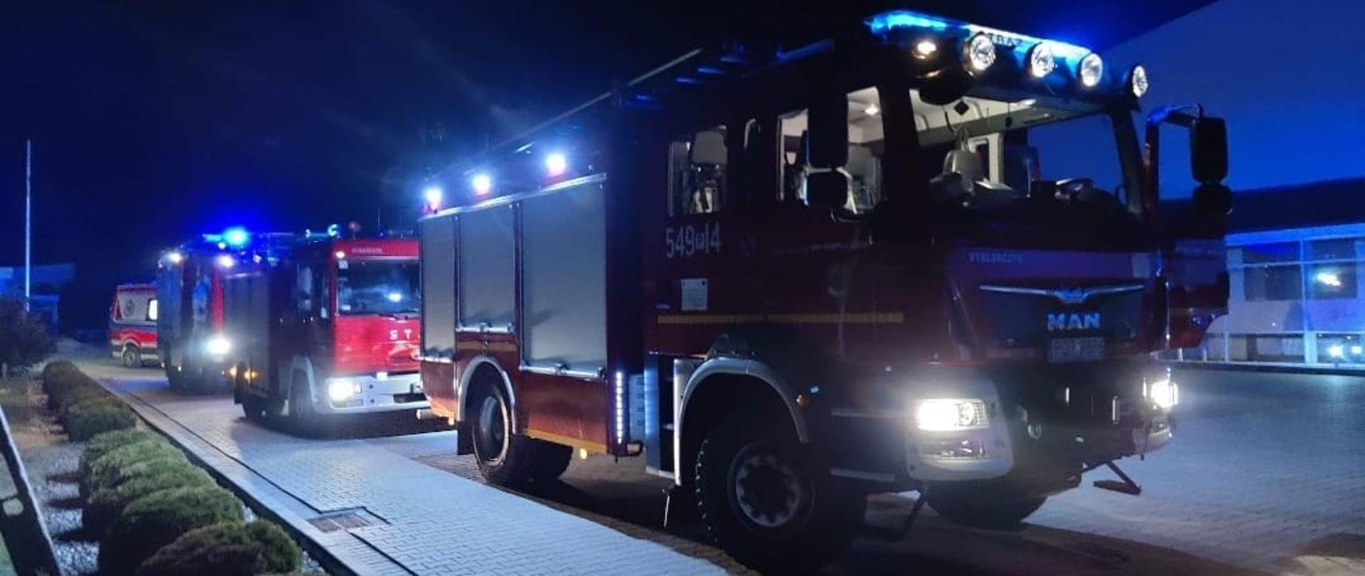 Na zdjęciu samochody gaśnicze stoją na pierwszym planie z uruchomioną sygnalizacją świetlną w trakcie akcji ratowniczej. Zdjęcie wykonane w porze nocnej. Na drugim planie karetka i budynek zakładu produkcyjnego.