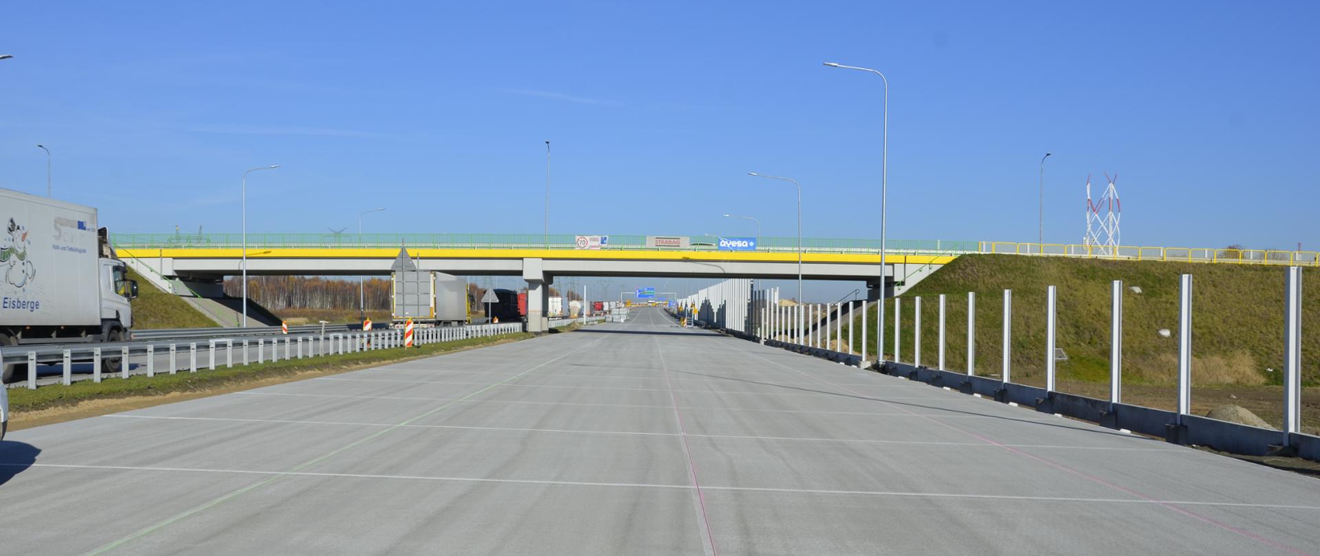 Wschodnia jezdnia autostrady A1 odc. C. W oddali na tle nieba widoczny wiadukt nad trasą w kolorach żółtym i zielonym. Wszystko oświetlone słońcem.