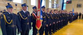 W hali sportowej w szeregu stoi grupa strażaków Komendy Powiatowej PSP w Ciechanowie. Na przedzie dowódca pododdziału, dalej sztandar i pozostali strażacy. 