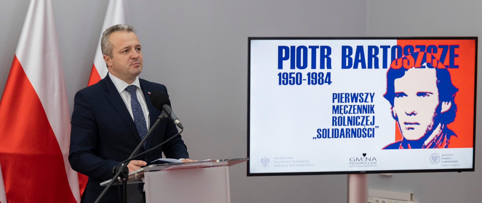 Jeden mężczyzna stojący przy mównicy i wypowiadający się. Obok telewizor z wyświetlonym plakatem