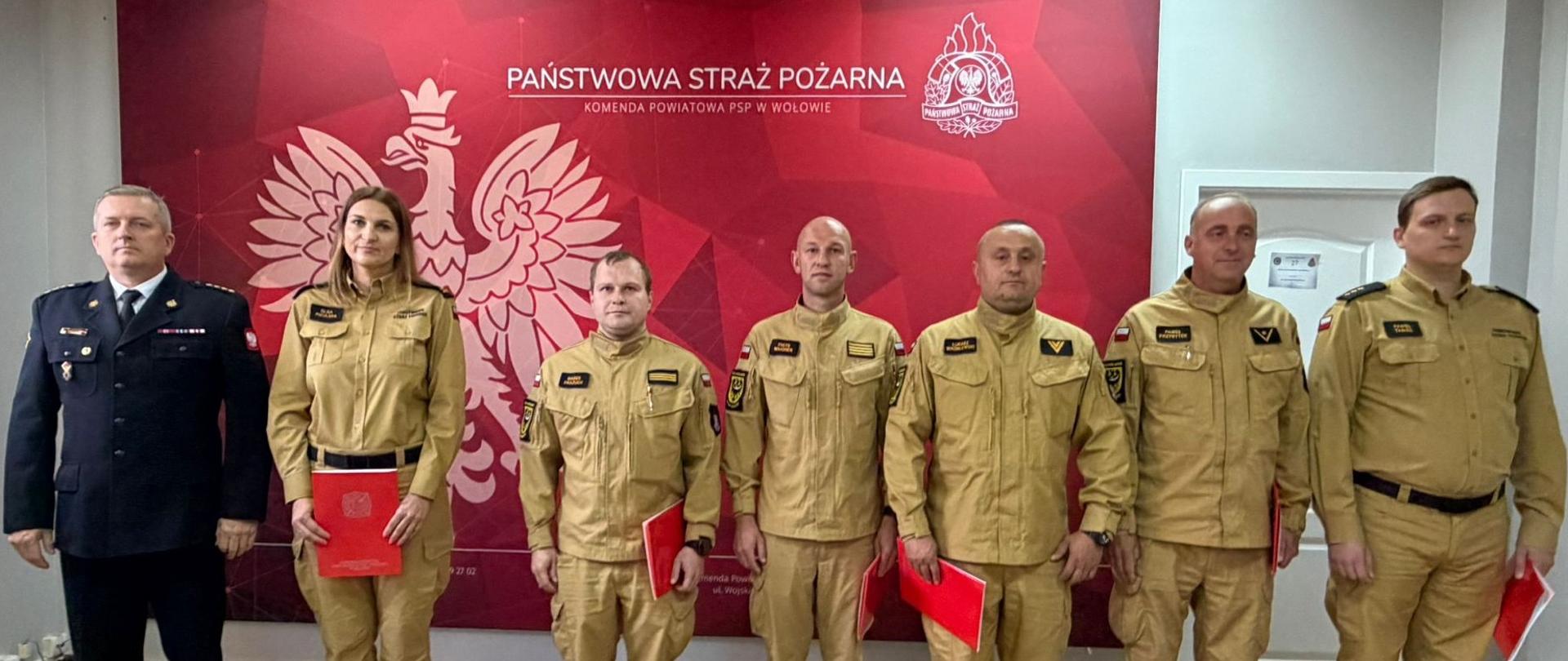 Siedmioosobowa grupa strażaków Państwowej Straży Pożarnej stoi w jednym rzędzie w sali Komendy Powiatowej PSP w Wołowie. Po lewej stronie stoi komendant powiatowy psp w ciemnogranatowym mundurze wyjściowym, a obok niego sześcioro strażaków w piaskowych mundurach służbowych, trzymających czerwone teczki.