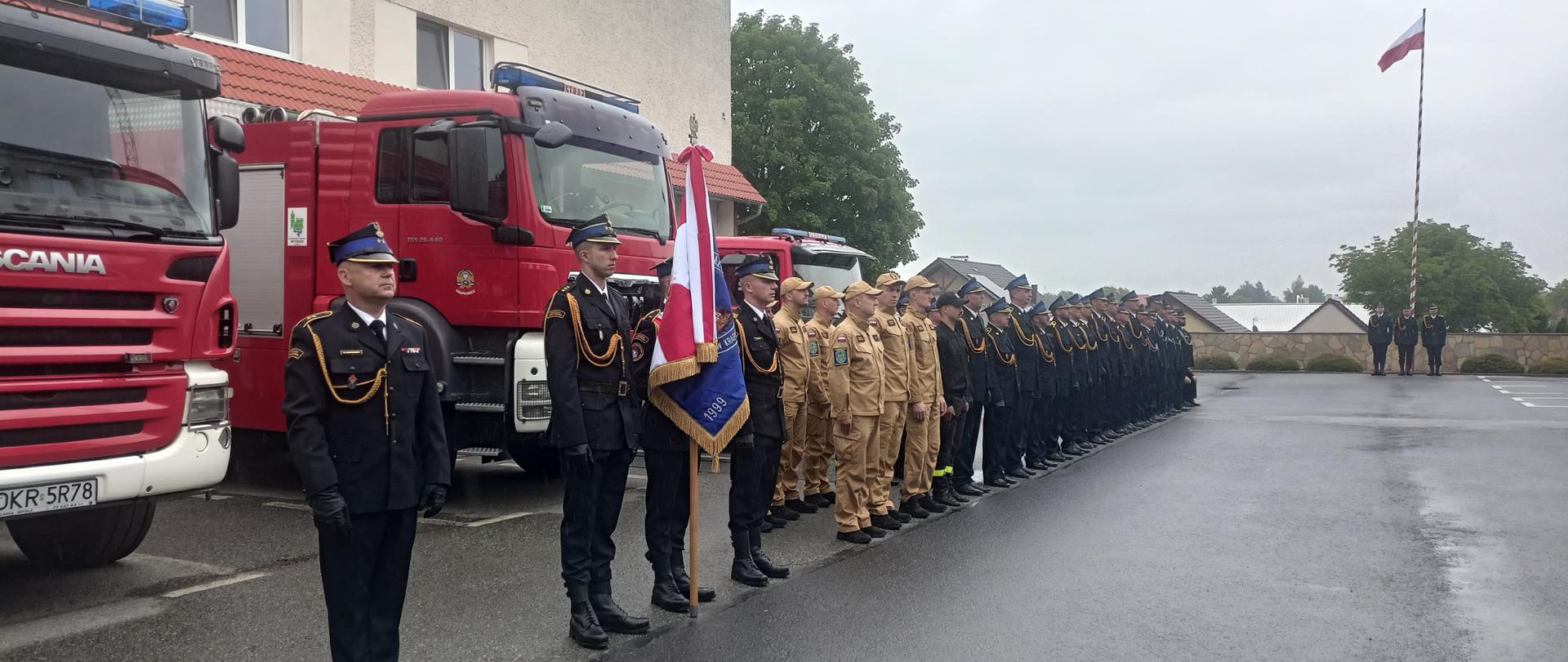 Na tle ustawionych przed budynkiem JRG samochodów pożarniczych ustawiony pododdział pożarniczy. Od lewej dowódca, następnie poczet sztandarowy, pododdział podziału bojowego w pisakowych mundurach służbowych, a dalej pozostali strażacy PSP i OSP w mundurach wyjściowych. Po prawej w oddali poczet flagowy ustawiony przy maszcie z powiewającą plaga RP. 