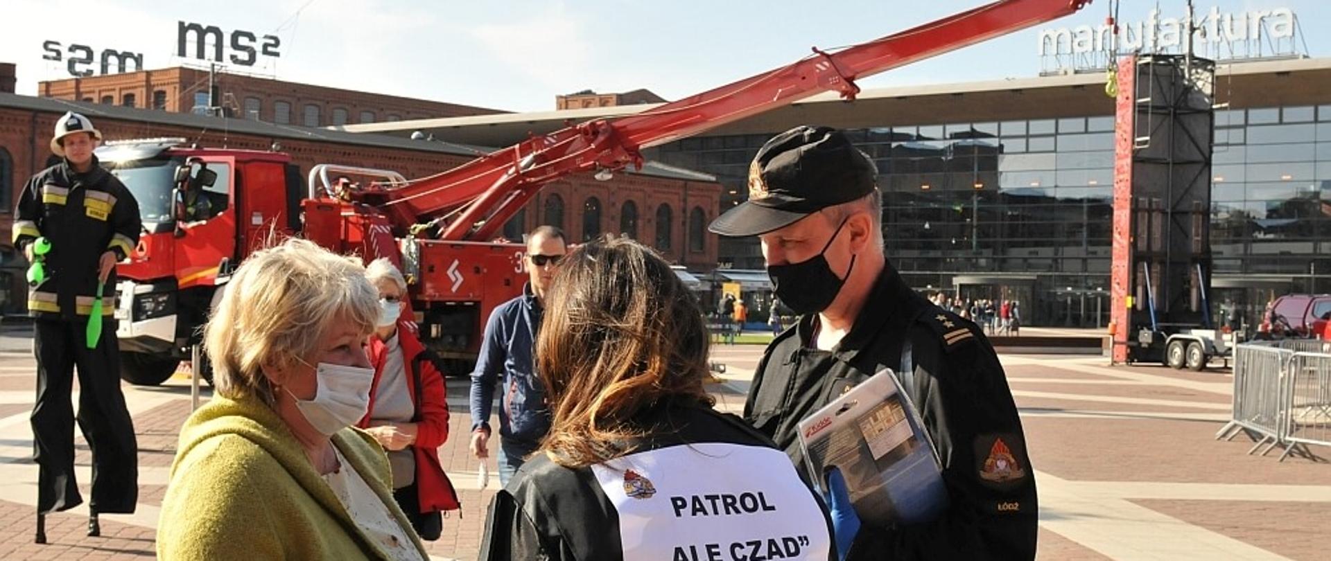 Dwoje funkcjonariuszy PSP w trakcie festynu "Czad i Ogień. Obudź czujność" w tle samochód ratownictwa drogowego oraz budynki centrum handlowego Manufaktura.