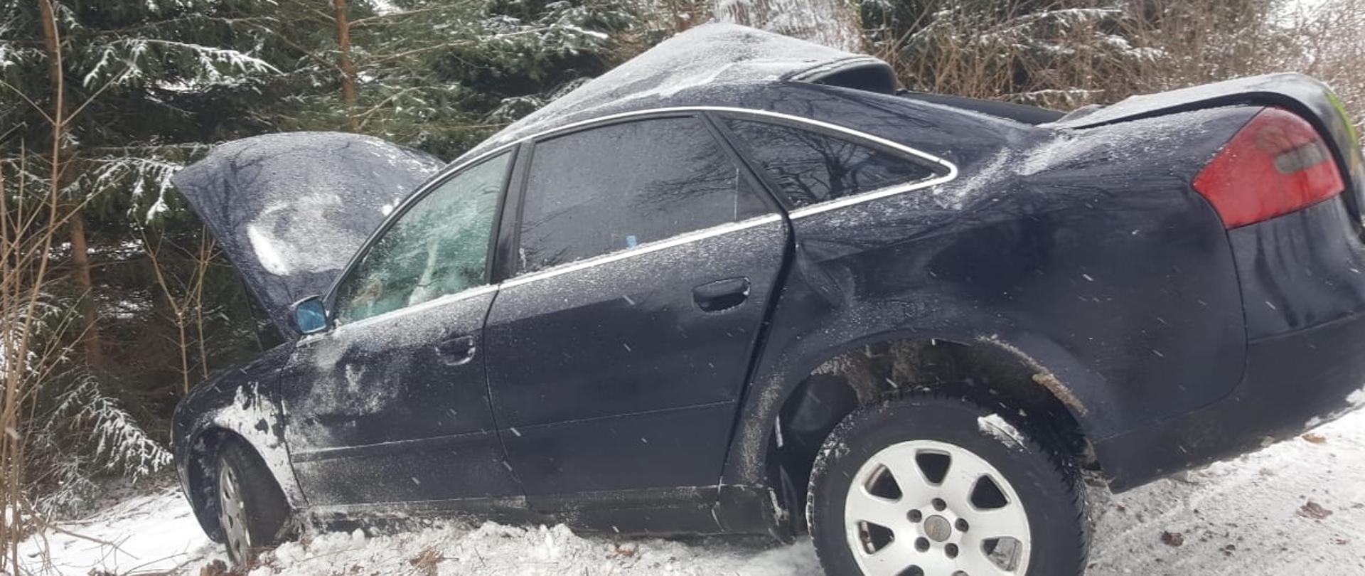 Ciemny samochód osoby uderzył lewym bokiem w drzewo i jest cały zniszczony. Dokoła leży śnieg. 