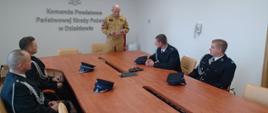 Komendant powiatowy Państwowej Straży Pożarnej w Działdowie stojąc odczytuje umowę. Czterech strażaków-ratowników OSP siedzi przy stole. Wszyscy ubrani w strażackie mundury.