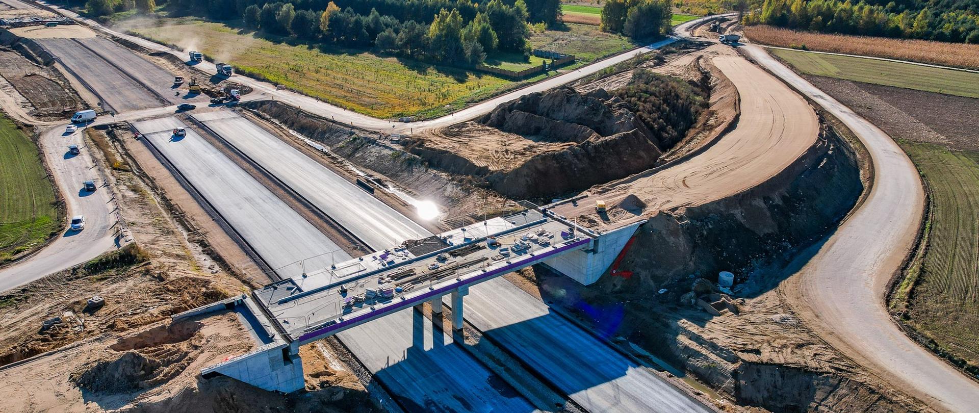 Widok z lotu ptaka na budowę drogi ekspresowej S19 w rejonie Malewice–Chlebczyn. Na zdjęciu widać szerokie pasy nowej jezdni w trakcie układania nawierzchni, most nad trasą z zamontowanymi barierami oraz otaczające nasypy ziemne i drogi techniczne. W tle lasy i pola.