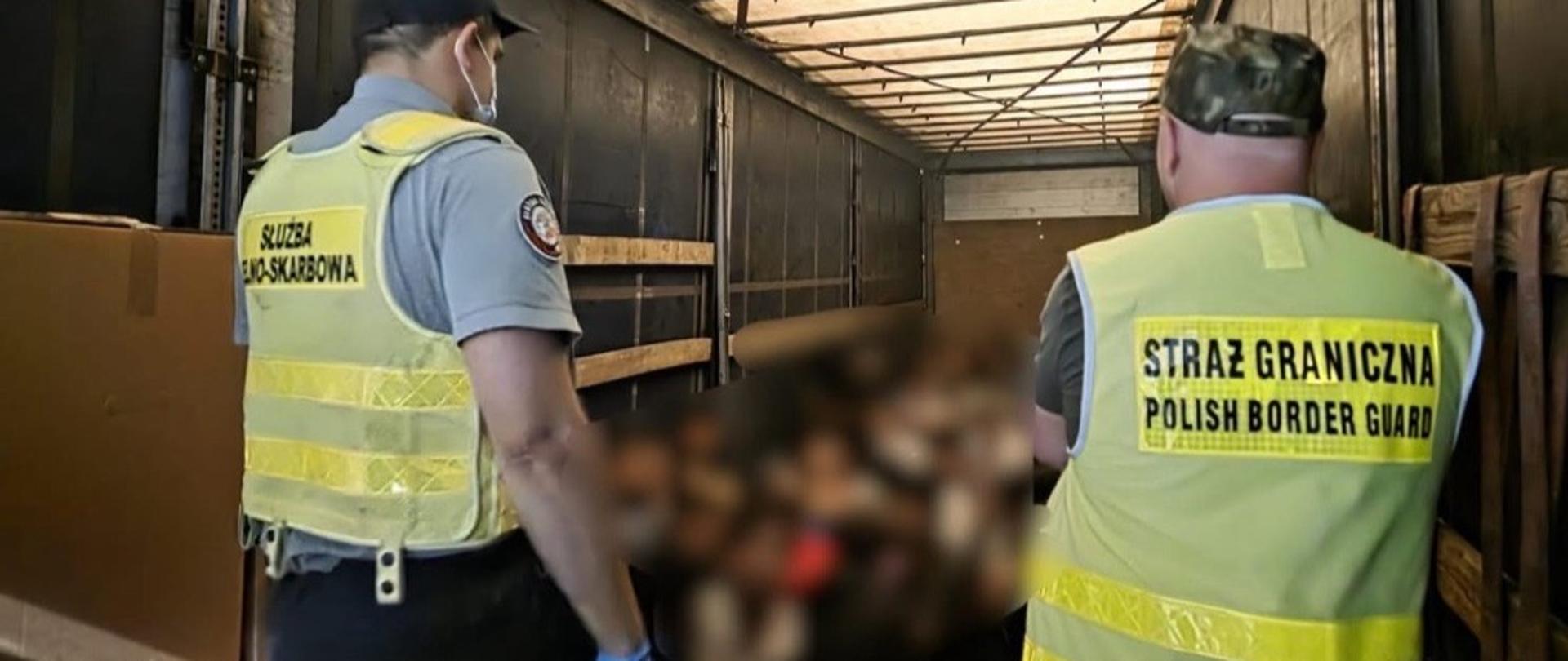 Funkcjonariusze Służby Celno-Skarbowej i Straży Granicznej stoją w naczepie ciężarowej obok kartonów; w głębi przestrzeni ładunkowej widoczne są siedzące osoby z zamazanym wizerunkiem.
