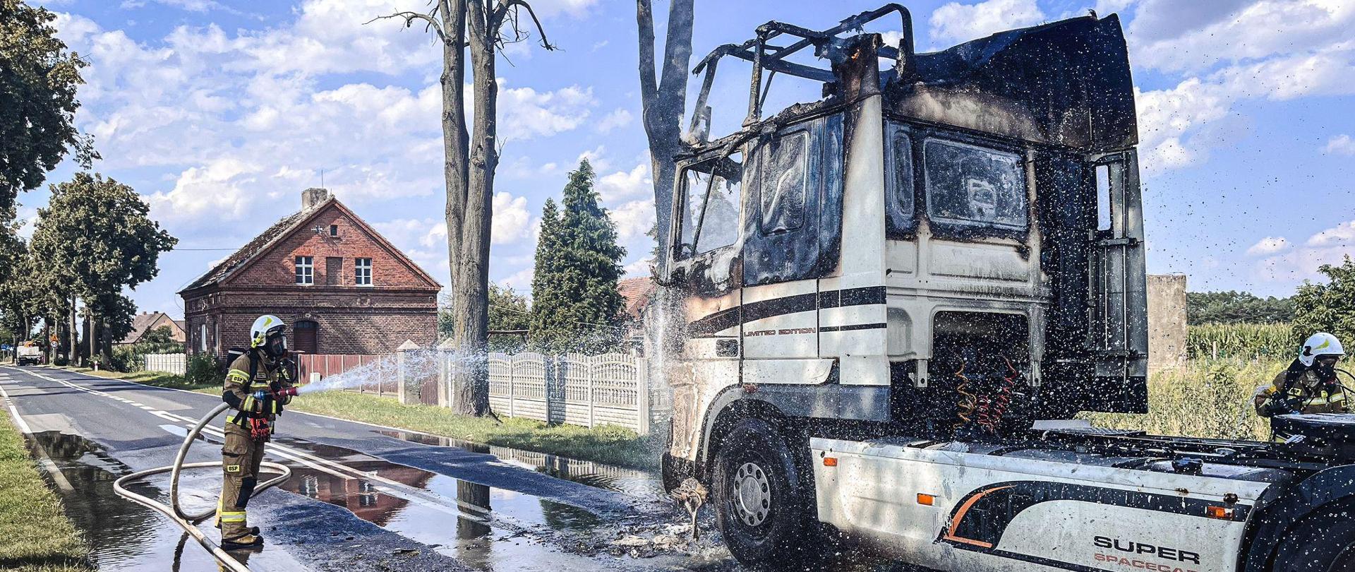 Strażacy zabezpieczeni w sprzęt ochrony dróg oddechowych dogaszają palący się pojazd. Pojazd z spaloną kabiną. 