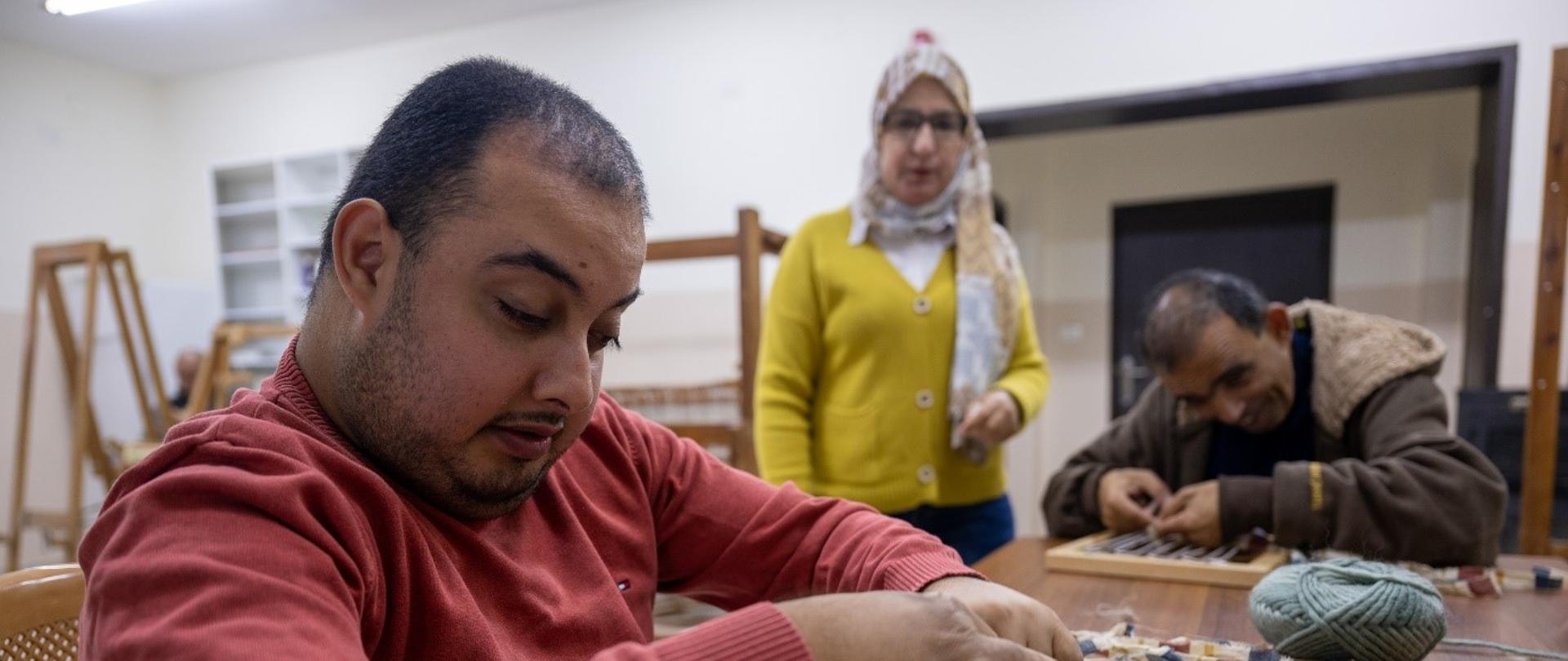Na pierwszym planie mężczyzna w czerwonej buzie siedzi przy stole i tka na małym krośnie. W tle drugi mężczyzna robi to samo. Kobieta stoi i przygląda się.