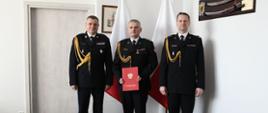 Trzech funkcjonariuszy Państwowej Straży Pożarnej stoi obok siebie za nimi stoją flagi Polski strażak w środku trzyma czerwoną teczkę.