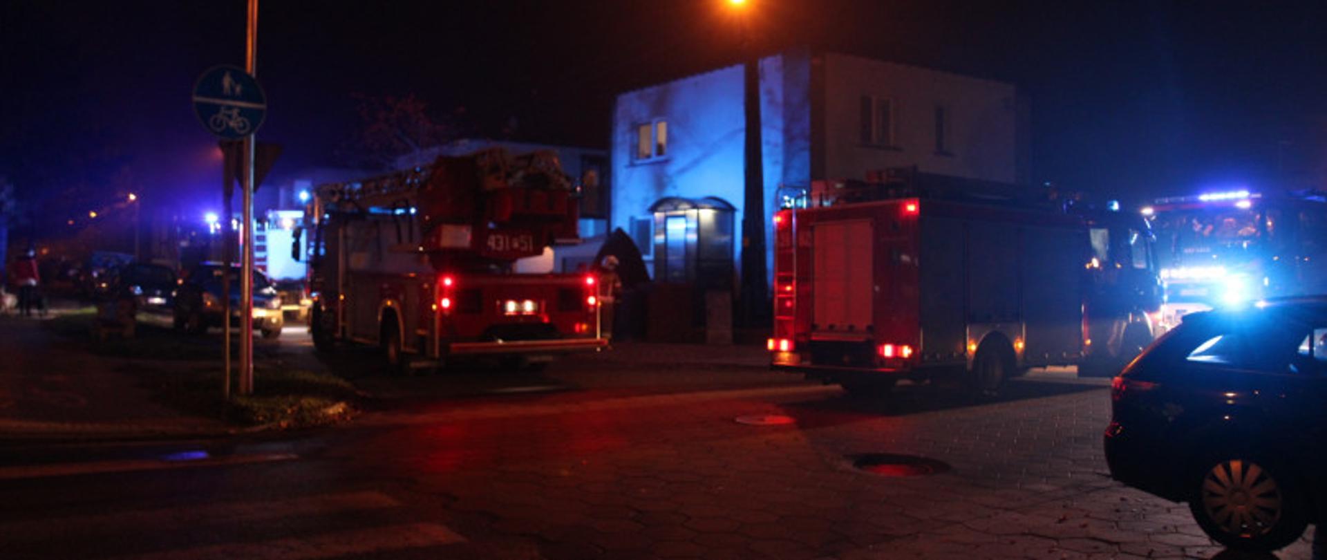 Zastępy straży pożarnej stoją przed budynkiem mieszkalnym. 