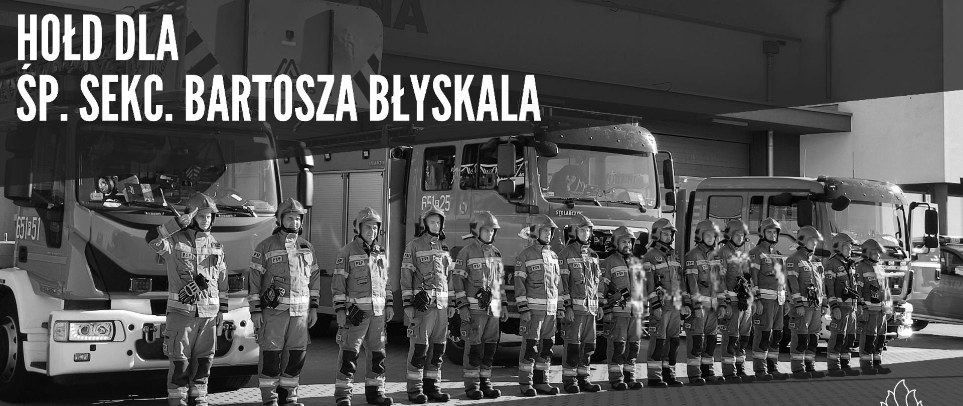 Zdjęcie przedstawia stojących na placu strażaków oddających hołd dla zmarłego strażaka.
W tle wozy strażackie i budynek Komendy.
