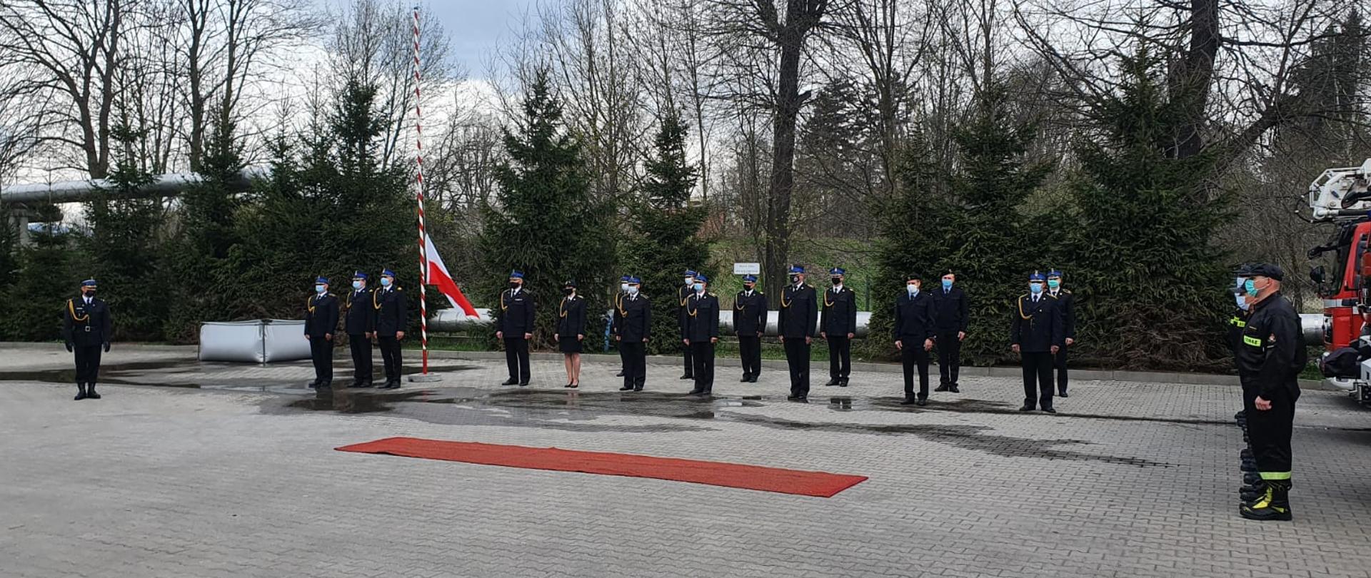 Apel z okazji Dnia Strażaka po lewej prowadzący uroczystość w tle widoczny poczet sztandarowy w składzie trójki funkcjonariuszy. po środku ustawieni strażacy w dystansie z uwagi na pandemie. Uroczystość odbywa się na placu Komendy Powiatowej Państwowej Straży Pożarnej w Kamiennej Górze. po prawej stronie zdjęcia dostrzegamy przód pojazdu pożarniczego - drabiny.