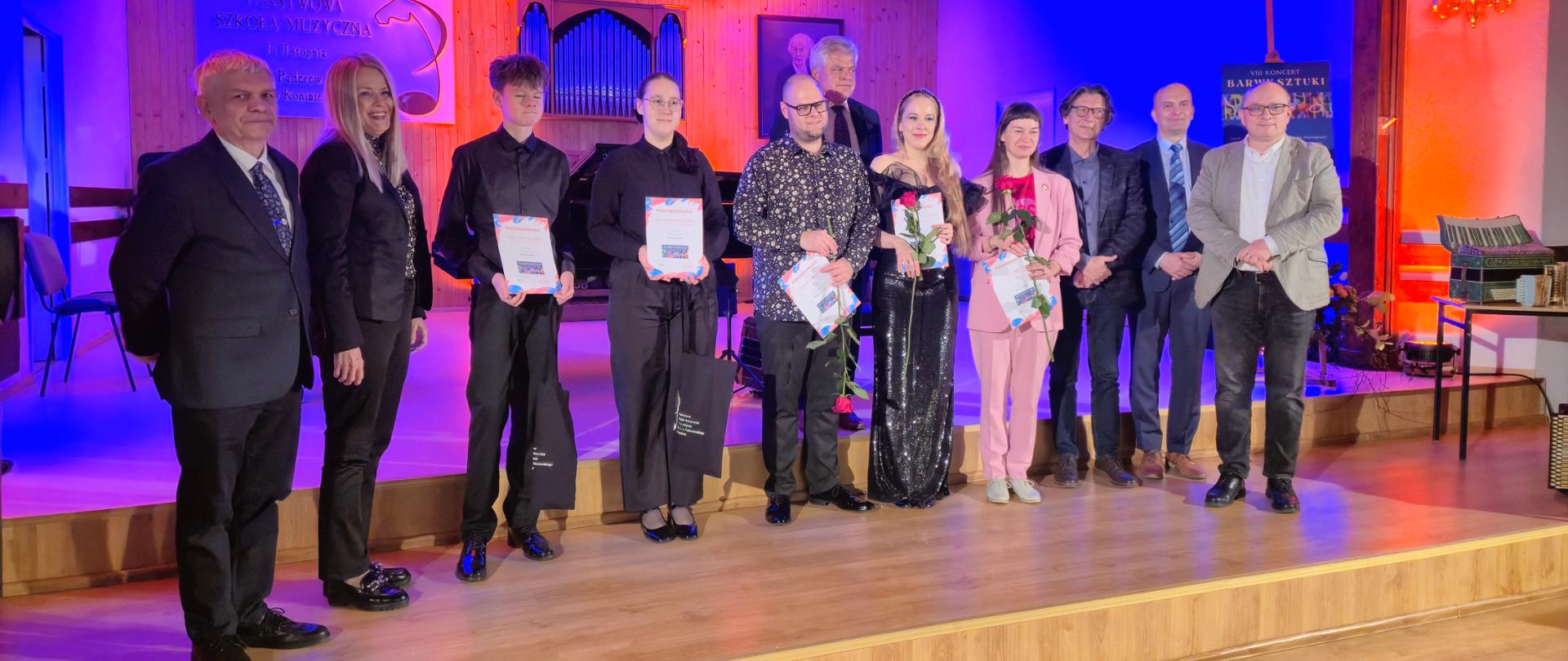 Grupa osób, w tym młodzi artyści trzymający dyplomy i kwiaty oraz przedstawiciele instytucji, pozuje wspólnie na scenie Państwowej Szkoły Muzycznej w Koninie po zakończonym występie artystycznym.